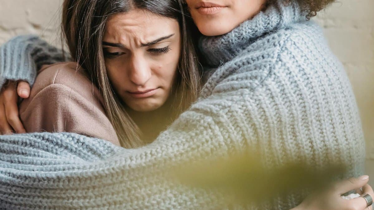 Anonymous woman in warn sweater comforting and embracing disappointed female friend near wall in room at home with green leaf