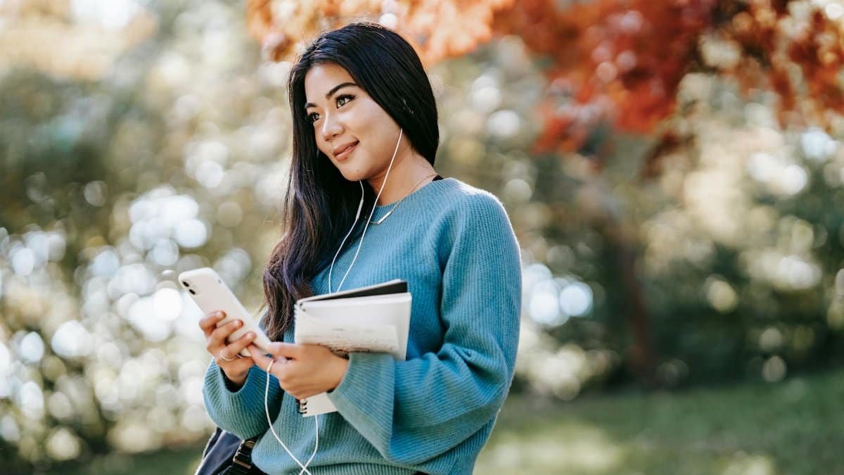 Cheerful young woman outdoors, using phone and earbuds, holding notebook, relaxing in a park.