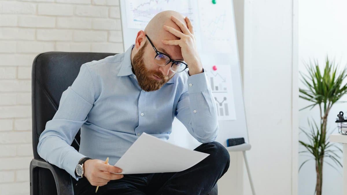 Bald man with glasses sitting in office looking worried at document.