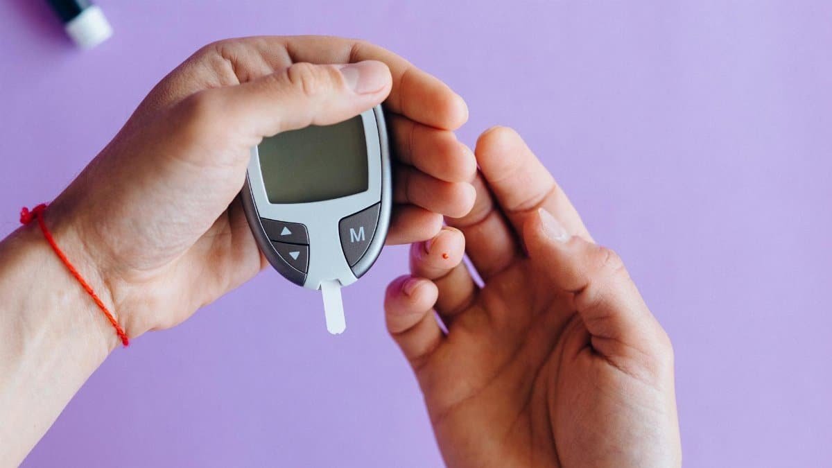 Hands holding a glucometer to test blood sugar levels on a purple background.