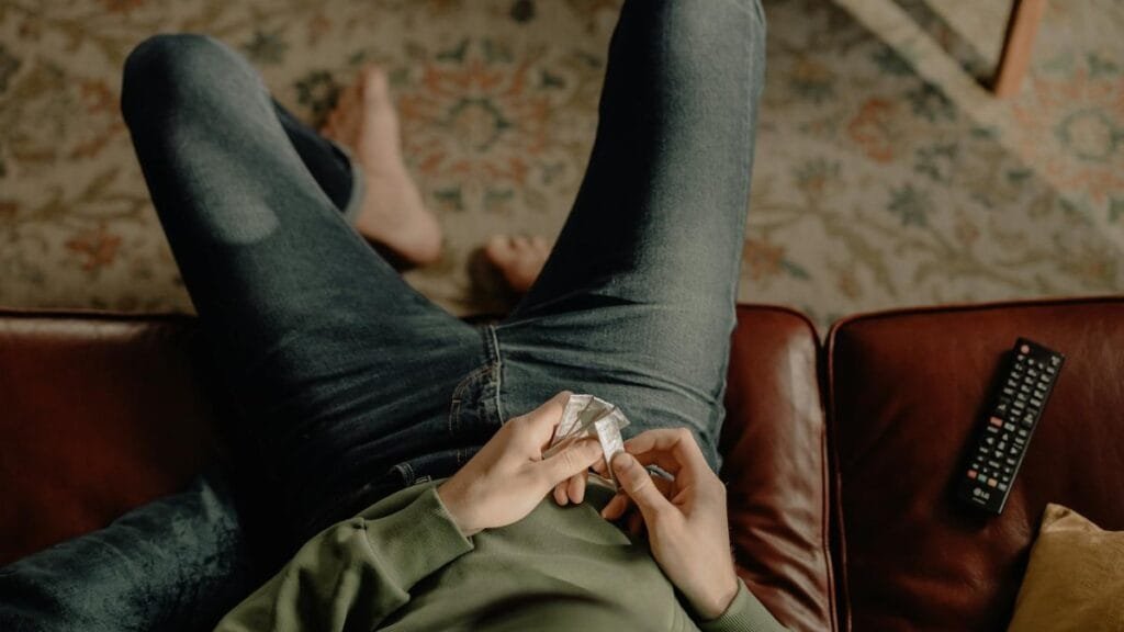 Person in hoodie relaxing on sofa with remote and chewing gum, cozy home vibe.