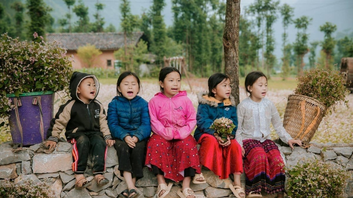 Five children sitting on a stone wall, wearing colorful jackets, outdoors with lush greenery.