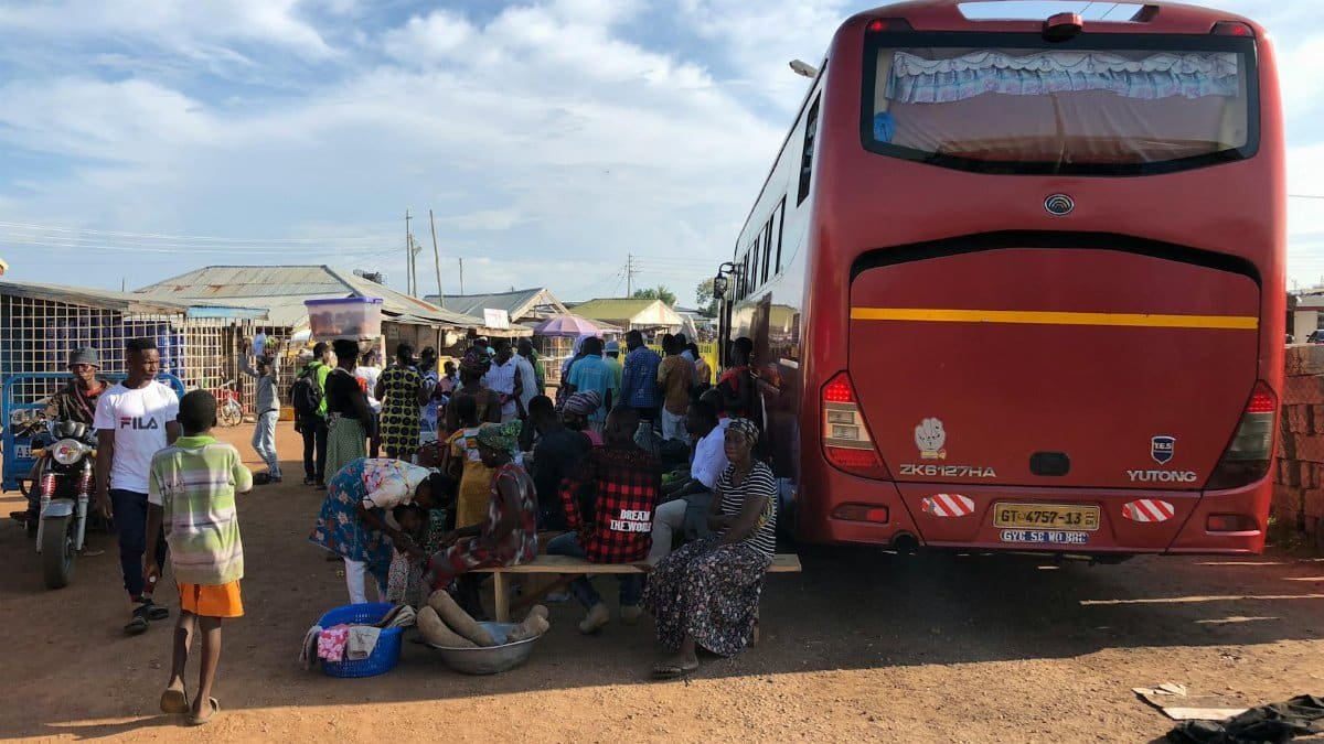 A busy scene at a Ghanaian bus station with locals gathered around a parked bus on a sunny day.