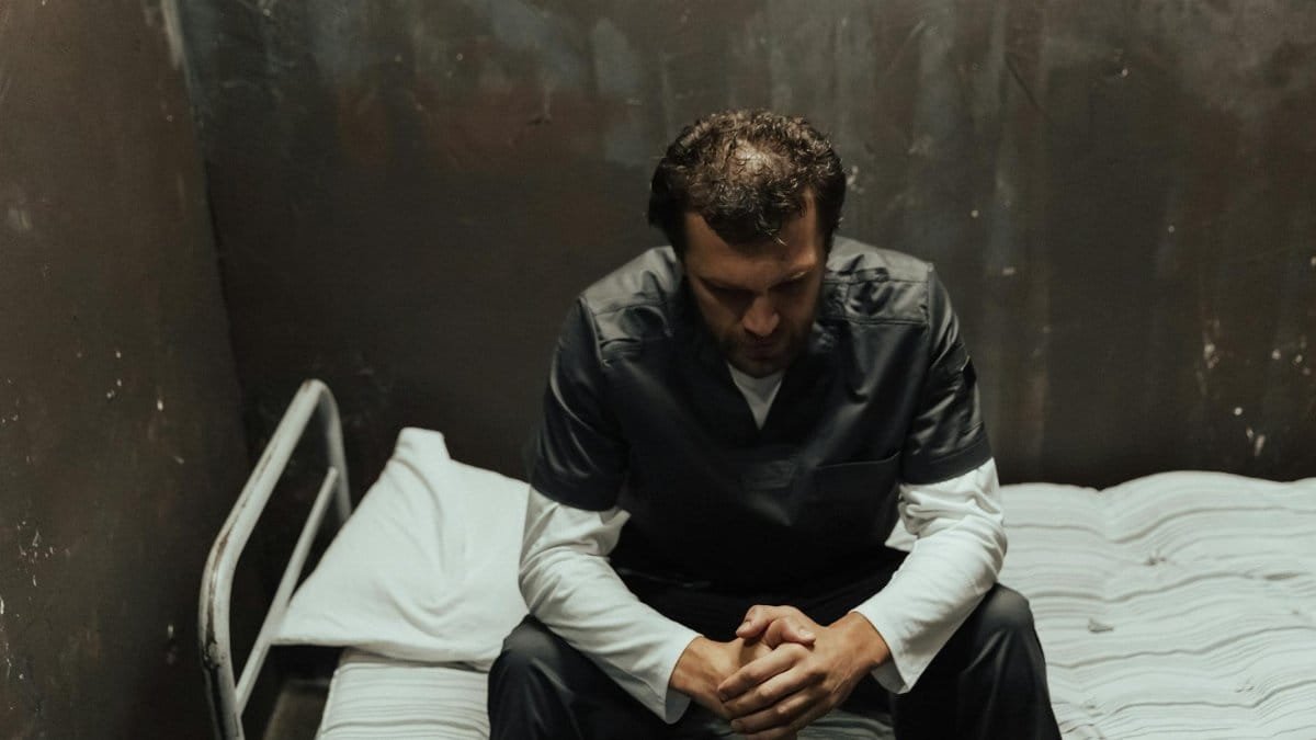 A male prisoner sits thoughtfully on the bed in a dimly lit cell, reflecting on life.