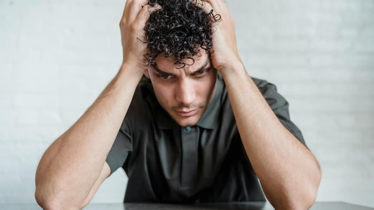 A young man sits indoors with his head in his hands, showing stress and worry.