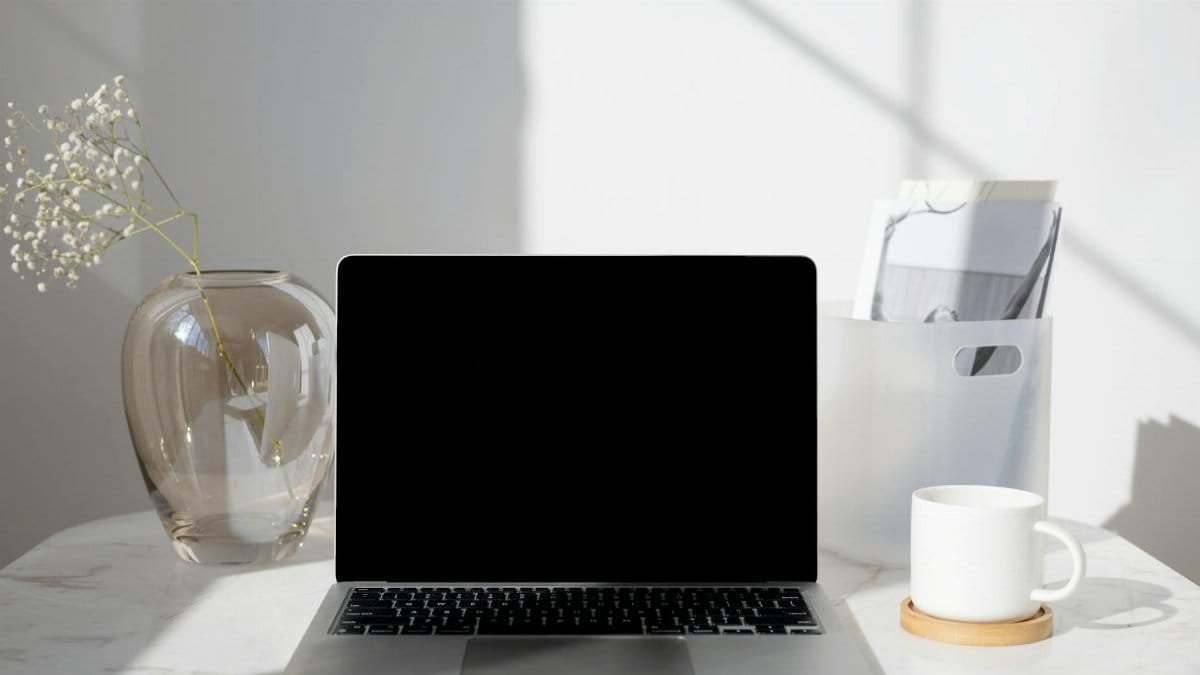 A clean and modern desk with a laptop, coffee cup, vase, and documents under bright daylight.