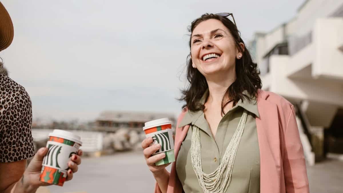 Smiling women enjoying conversation and coffee outdoors on a sunny day, promoting connection and lifestyle.
