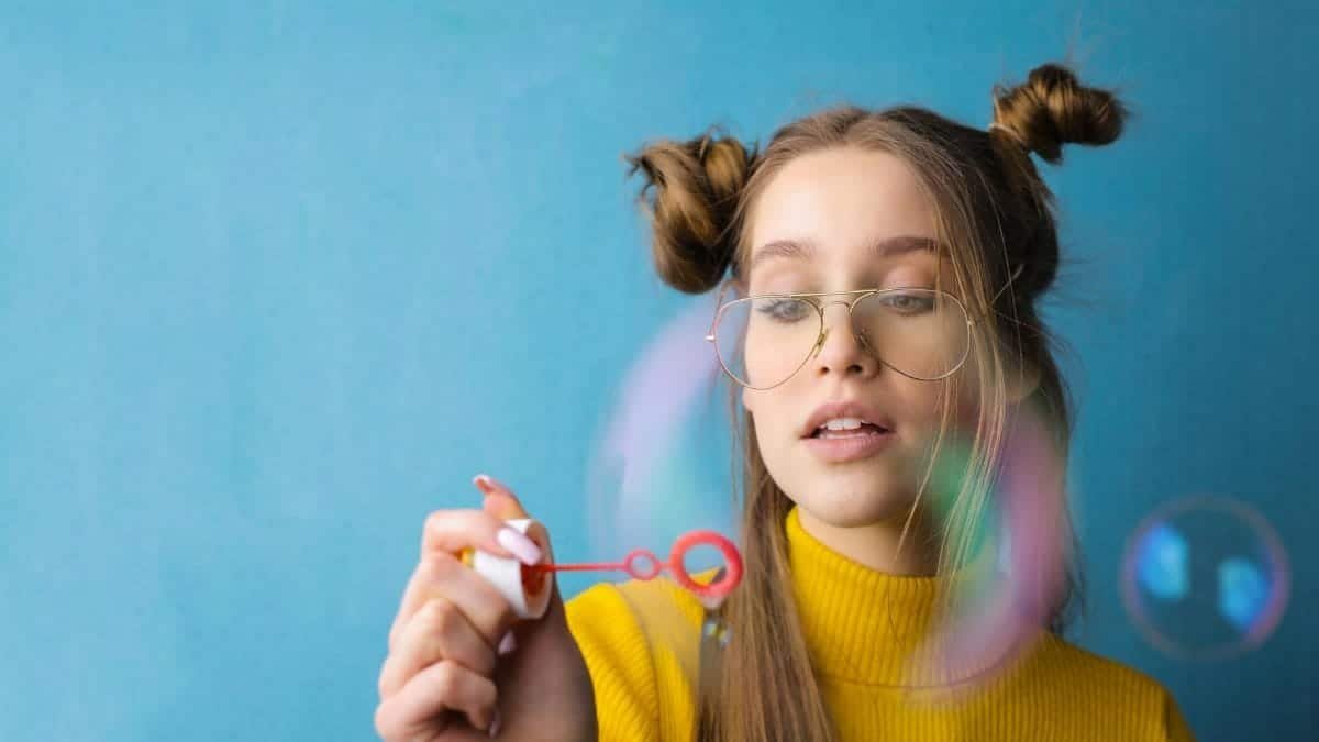 A young woman in glasses and a yellow sweater blowing bubbles with an amused expression.