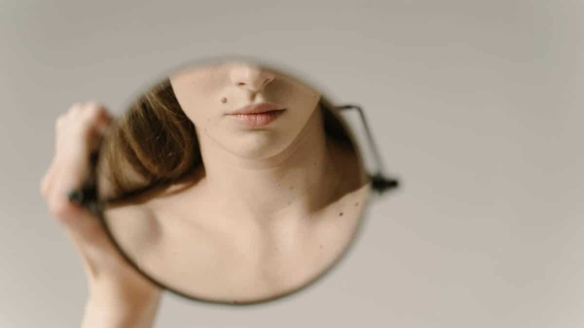 A close-up reflection of a woman's face in a handheld mirror, focusing on her lips and beauty mark.