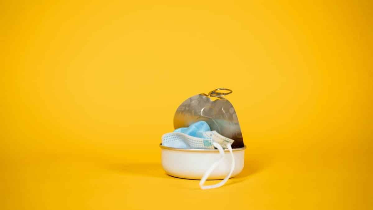 Close-up of a face mask in an open tin can against a bright yellow background, symbolizing pollution and waste.