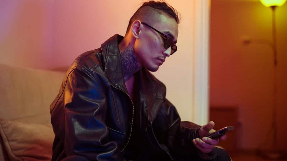 Trendy man with neck tattoos and sunglasses using smartphone on a sofa indoors.