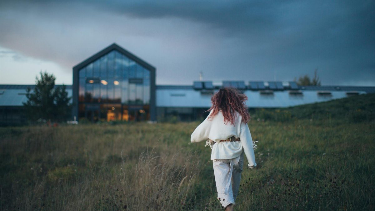 A woman walks through a grassy field towards a modern building during twilight, creating a serene and contemplative scene.