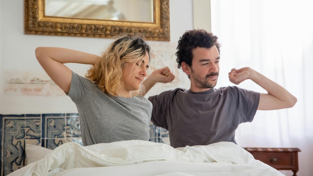 A couple begins their morning stretching in bed with soft daylight filtering through curtains.