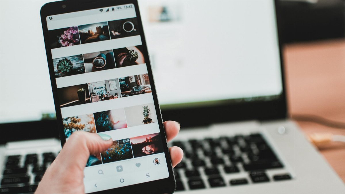 A person uses a smartphone to browse social media in front of a laptop, emphasizing modern technology and lifestyle.