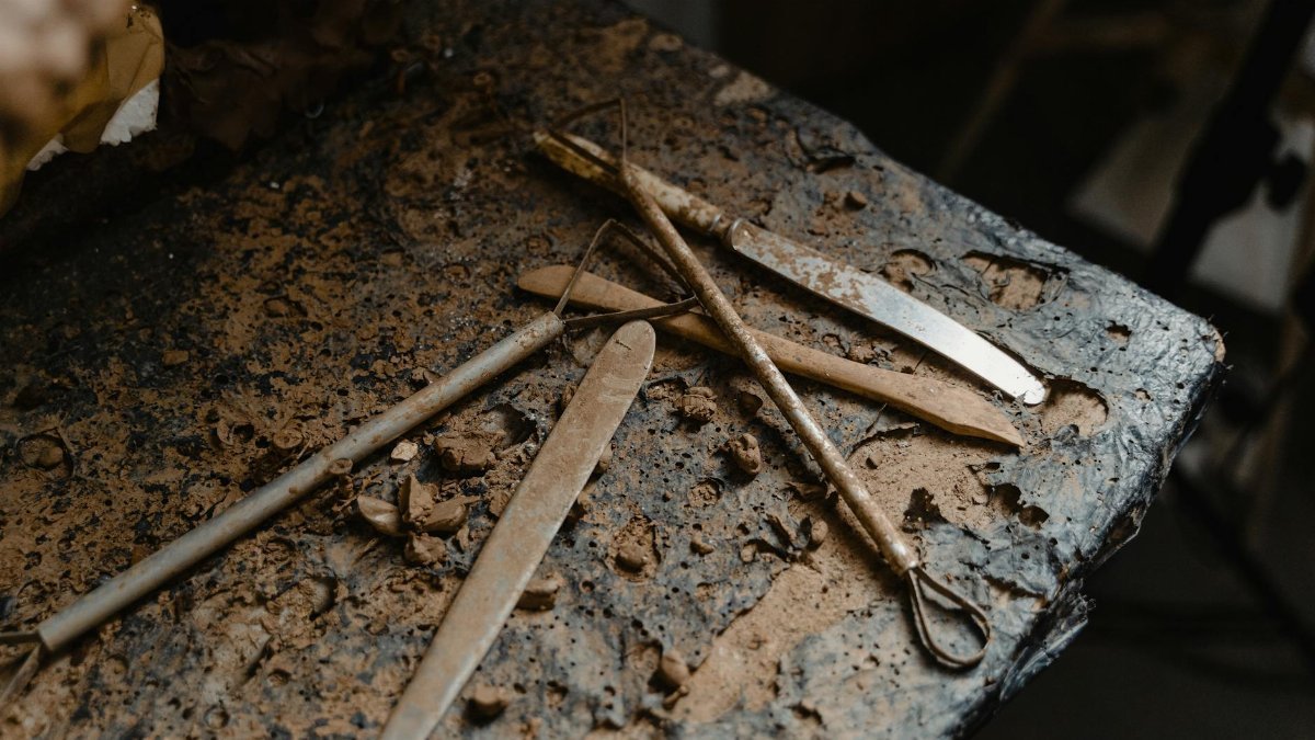 Close-up of rusty sculpture tools laid out on a muddy workbench in a dimly lit studio.