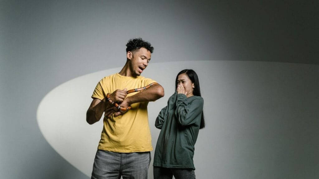 A man holds a snake while a woman appears scared, depicting a moment of fear and anxiety.