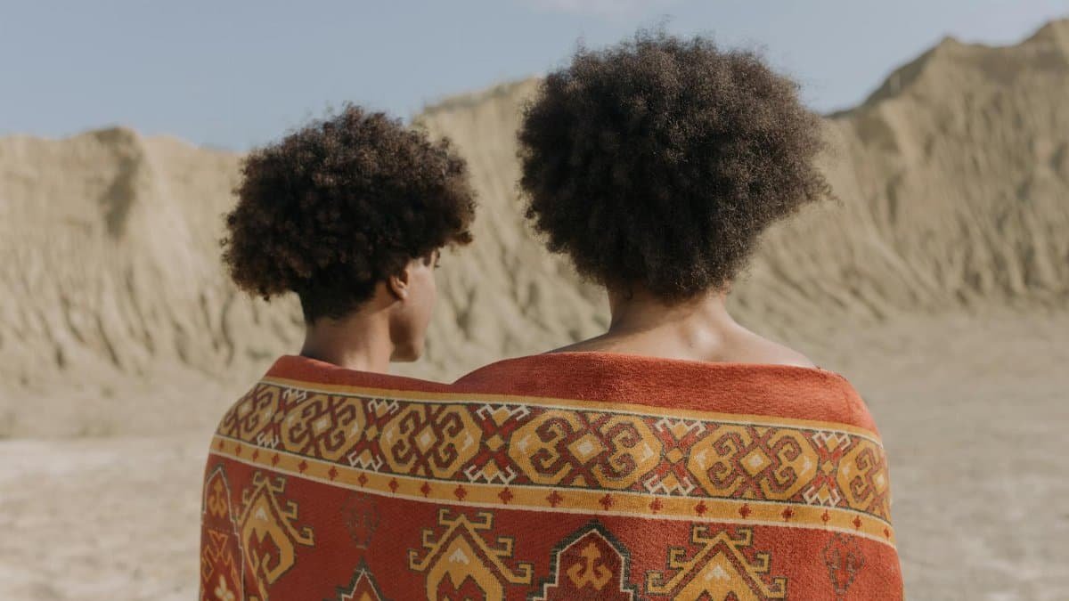 Two people with afro hair wrapped in a colorful shawl stand in an arid desert landscape.