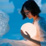 A woman in a dream-like setting holding a glowing sphere surrounded by cloud-like formations.