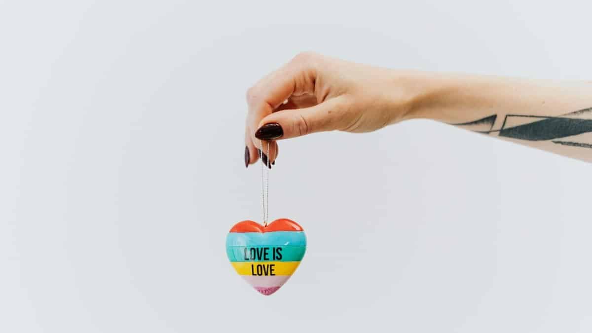 A tattooed hand holds a colorful heart keychain with 'Love is Love' text, symbolizing LGBTQ pride and acceptance.