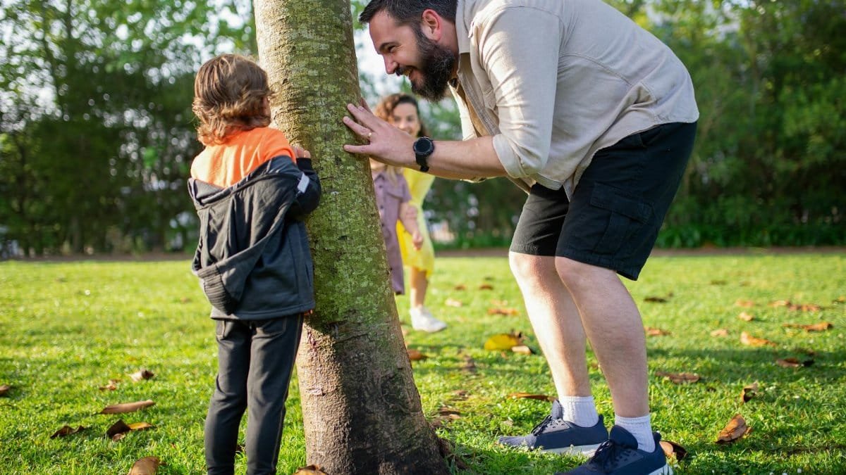 A father and children enjoy playing hide and seek around a tree in a sunny park.