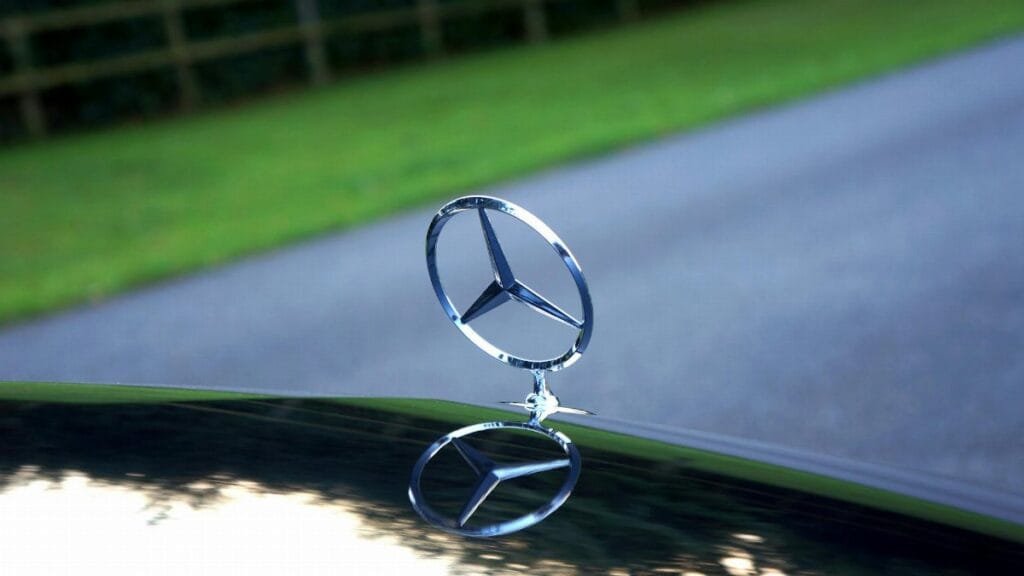Elegant close-up of a Mercedes-Benz emblem reflecting on a car hood with a blurred road and greenery in the background.