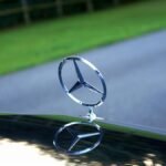 Elegant close-up of a Mercedes-Benz emblem reflecting on a car hood with a blurred road and greenery in the background.