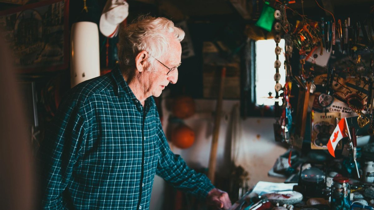 Senior man in a checked shirt exploring a cluttered indoor space with personal items and Canadian flag.