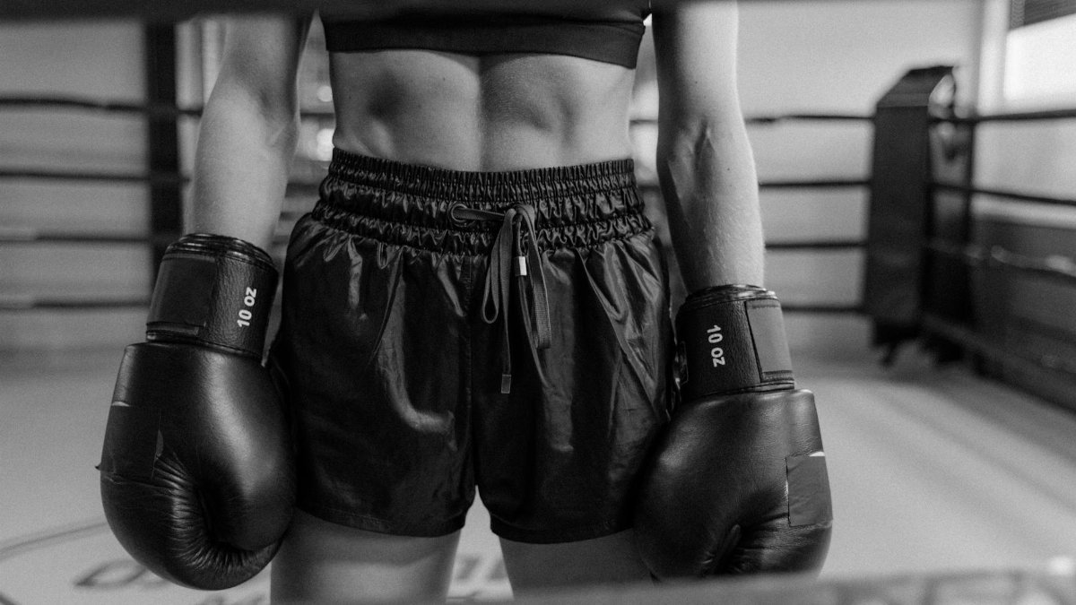 Black and white photo of a boxer in a gym wearing gloves and shorts, showcasing fitness.