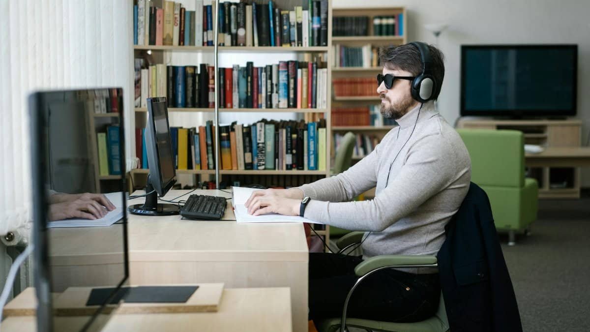 A blind man uses a computer with headphones in a library, highlighting technology's role in accessibility.