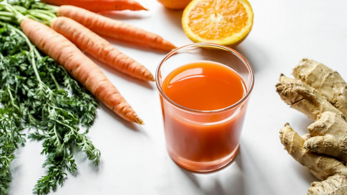 Fresh carrot ginger and orange juice surrounded by ingredients on a white background.