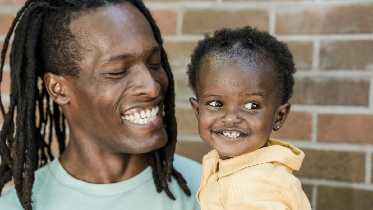 A father with dreadlocks joyfully holding his smiling young daughter against a brick wall backdrop.