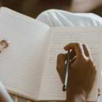 Close-up of hands writing in a journal with a pencil on a seated lap.