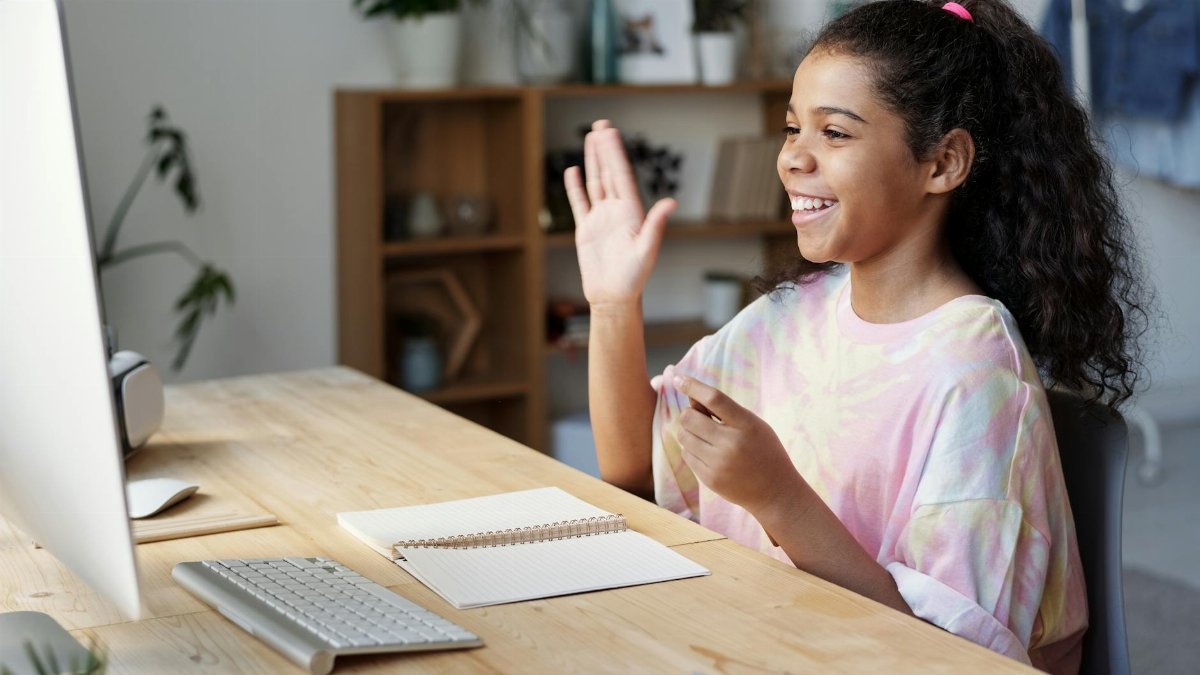 A happy girl waves during an online class at home, sitting at a desk with a computer.