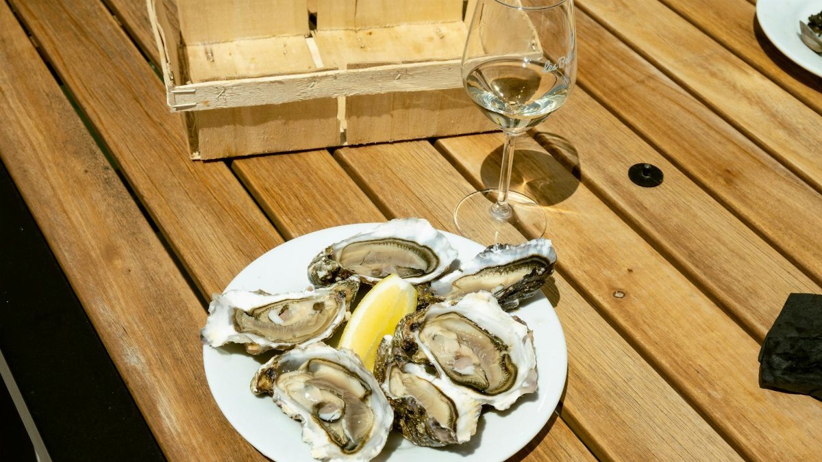 Plate of fresh oysters with a lemon wedge and wine glass on a wooden table outdoors.