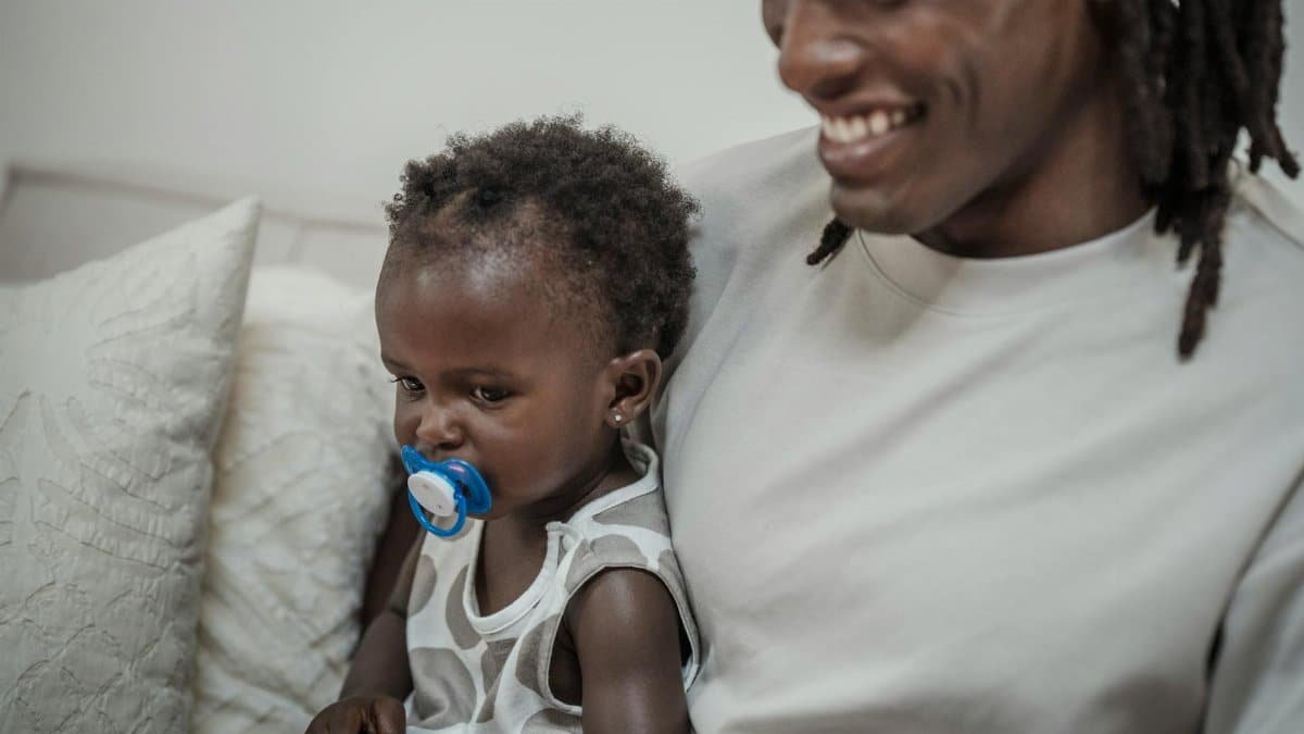A smiling father holding his daughter with a soother, sitting together indoors.