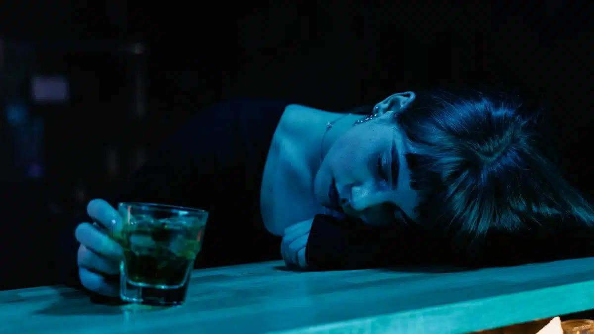 A woman resting her head on a bar table while holding a drink, in a moody, dimly-lit nightclub setting.