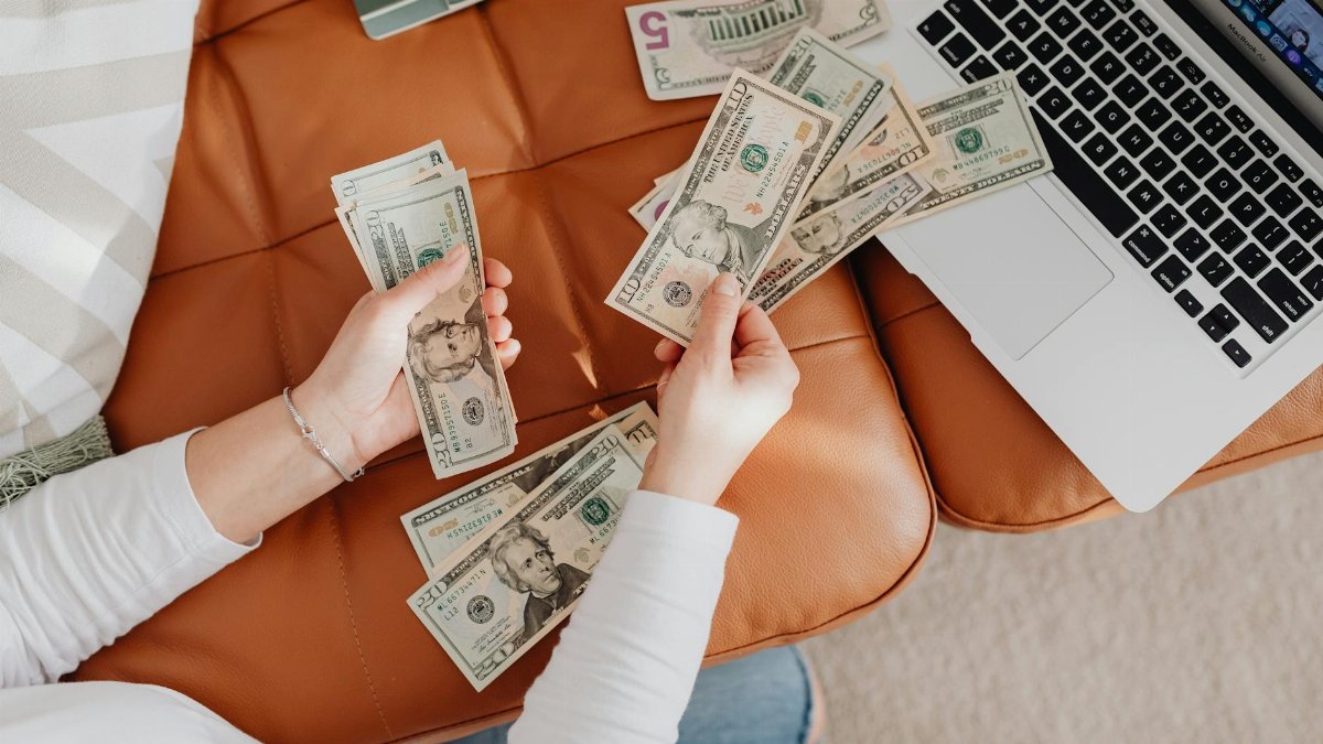 A person counts US dollar bills on a couch with a laptop, symbolizing financial planning.