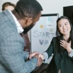 A diverse business team in formal attire exchanging a handshake in a modern office setting, signifying cooperation.