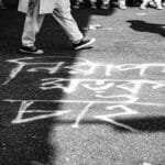 Monochrome photo capturing people walking over chalk writing on a city pavement.