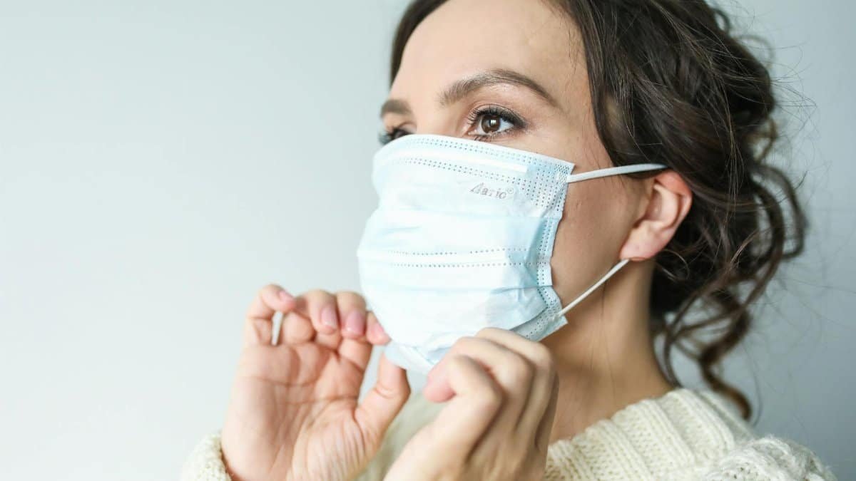 Adult woman adjusting blue surgical mask indoors for health safety.