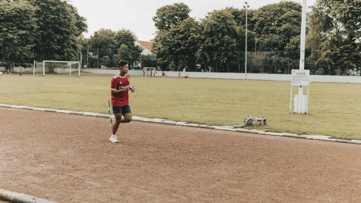 A young man in athletic attire jogging on an outdoor track in a park setting.