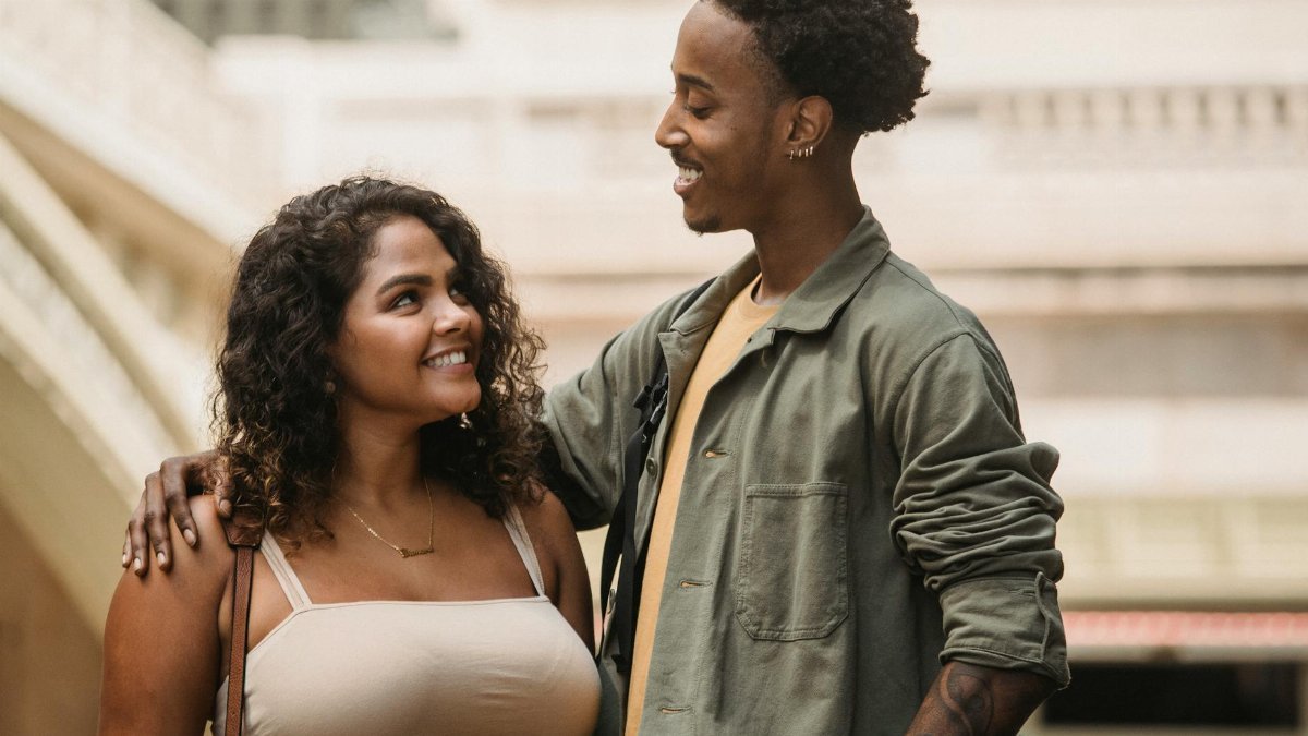 Positive multiracial young couple wearing casual clothes standing and hugging while looking at each other on blurred city street in daylight