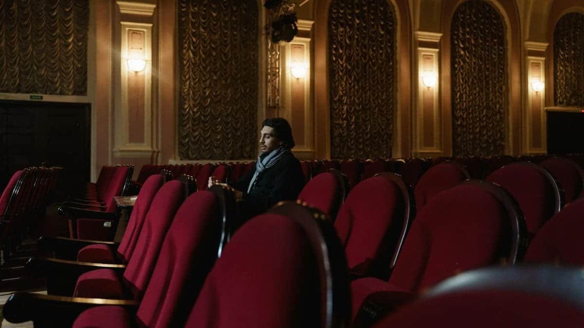 A man sits alone in an opulent theater, surrounded by red chairs and ornate decor.
