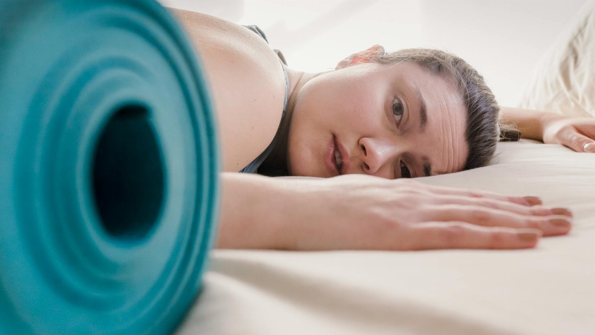 A tired woman rests on a bed beside a yoga mat, capturing post-exercise fatigue.