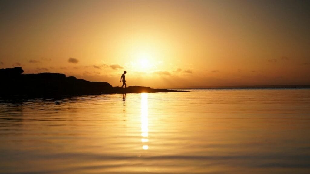 A lone silhouette walks along Currarong Beach at sunset, reflecting on serene waters.
