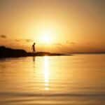 A lone silhouette walks along Currarong Beach at sunset, reflecting on serene waters.