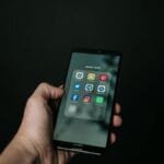 Close-up of a hand holding a smartphone displaying various social media app icons on a dark background.