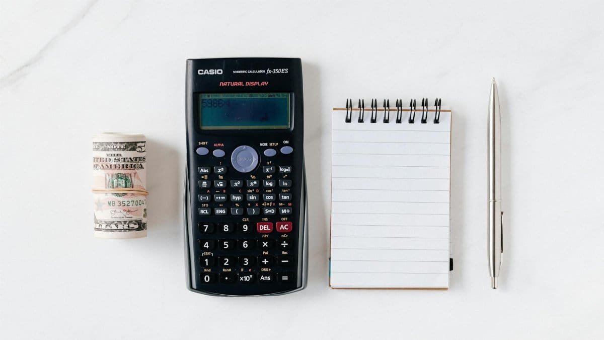 Top view of financial tools including a calculator, notepad, pen, and rolled cash on a white background.