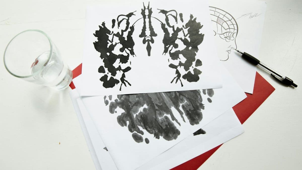 An artistic arrangement of inkblot test sheets with a pen and glass on a table.