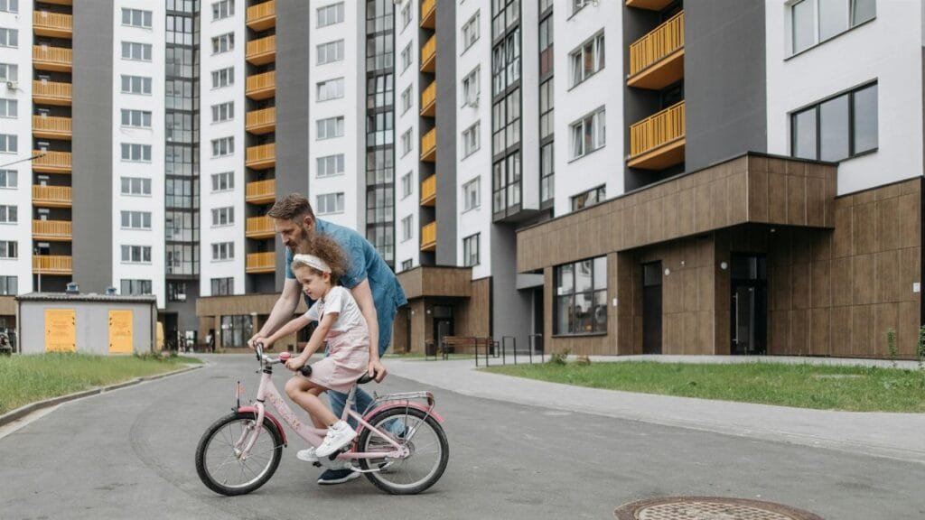 Father guiding daughter on a bike ride outside modern apartments. Bonding and teaching moment.